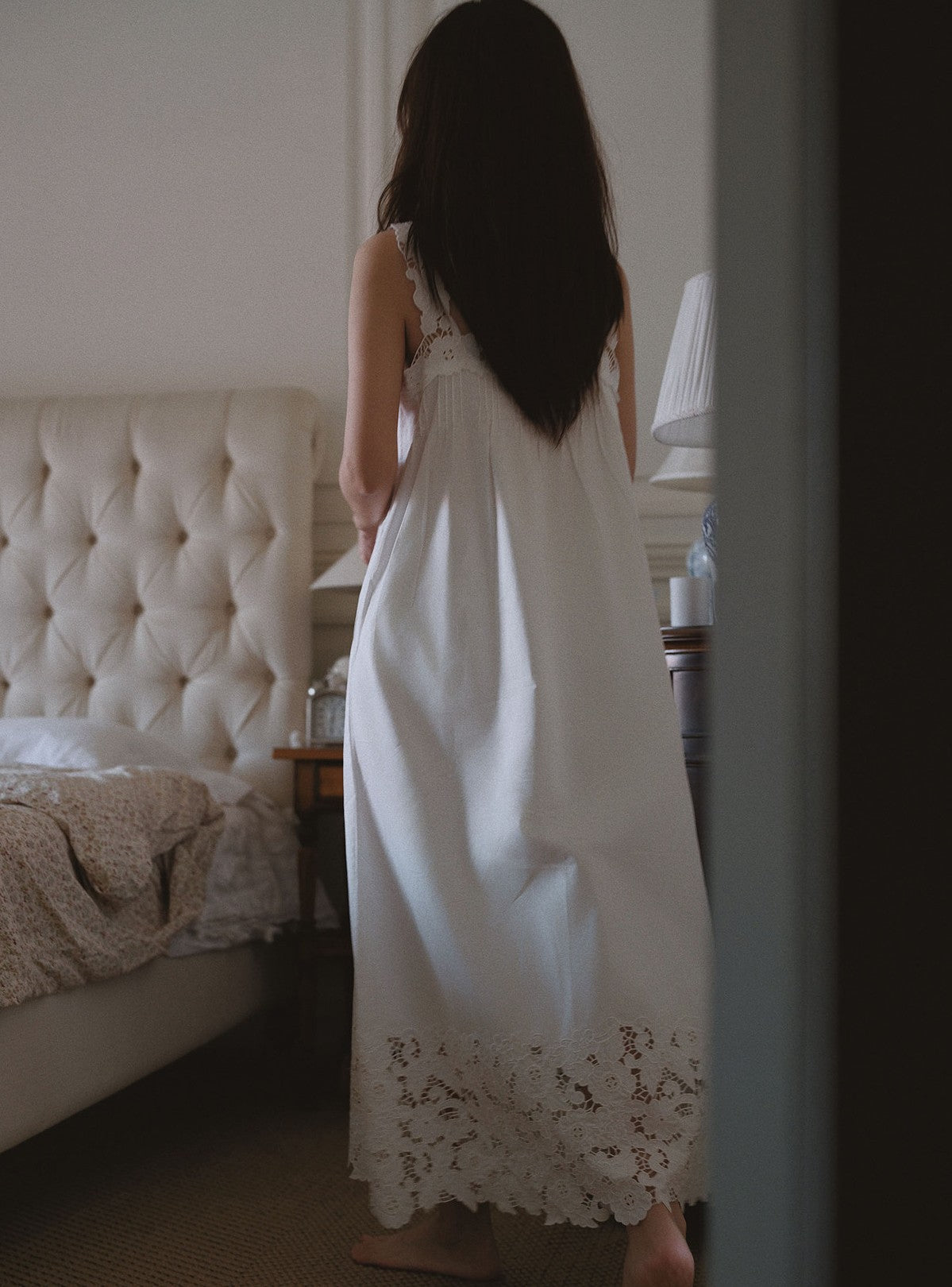 A woman with long dark hair stands with her back to the camera in a bedroom, wearing a white sleeveless nightgown with lace trim at the hem.