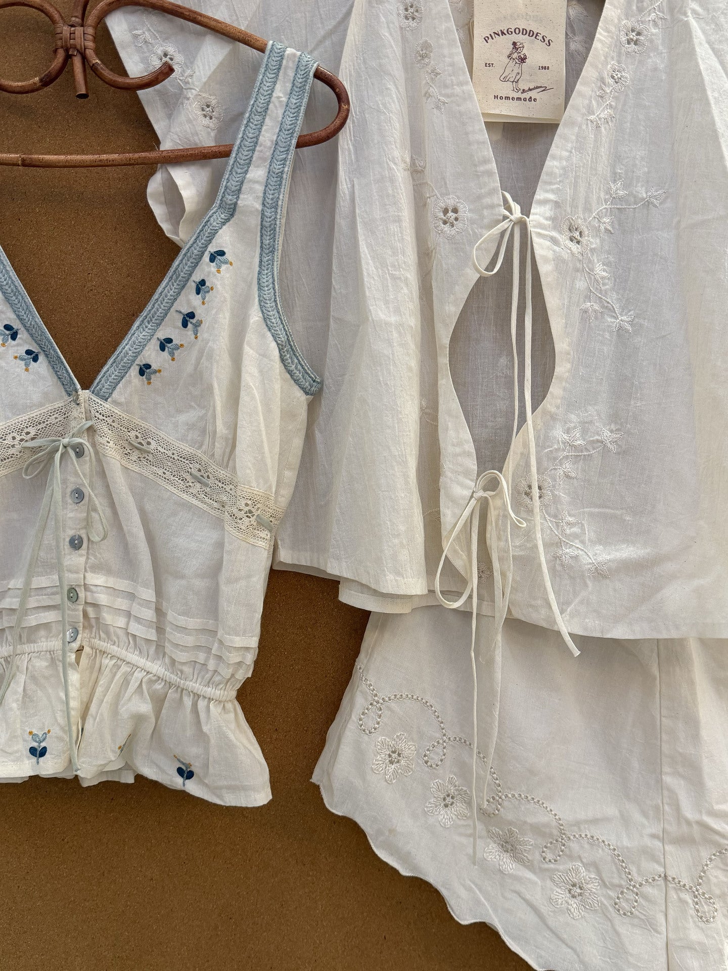 A white embroidered two-piece set consisting of a sleeveless top with light blue trim and a matching short-sleeve blouse, both hanging on a wooden hanger against a brown background. The brand label 'Peachy Sweets' is visible.