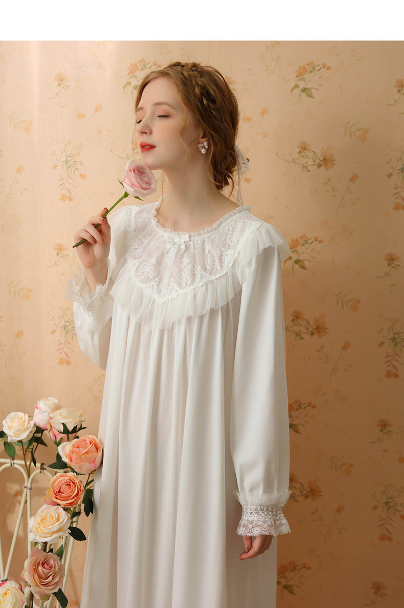 A woman wearing a long white nightgown with lace trim and ruffled details, holding a pink rose against a warm floral wallpaper background.