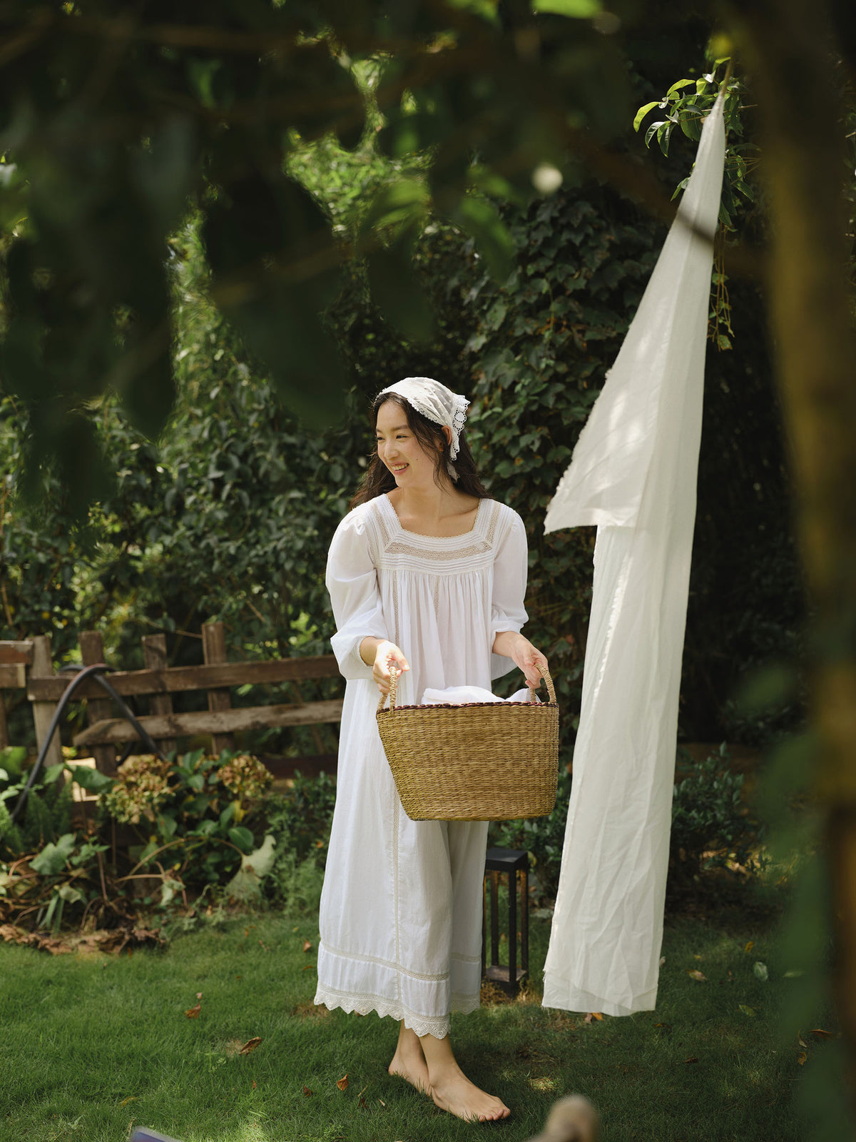 A woman stands barefoot on a grassy garden lawn wearing a long white nightgown and holding a woven basket, with a white sheet hanging from a tree nearby in a lush green outdoor setting.