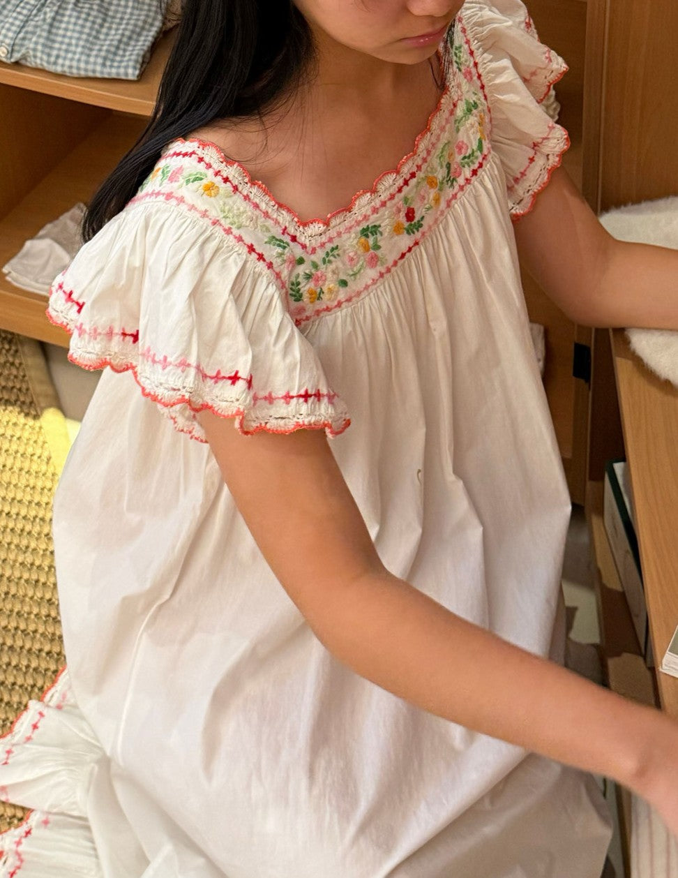 A girl wearing a white cotton nightgown with colorful floral embroidery and ruffled trim on the neckline and sleeves, shown in a warm indoor setting.