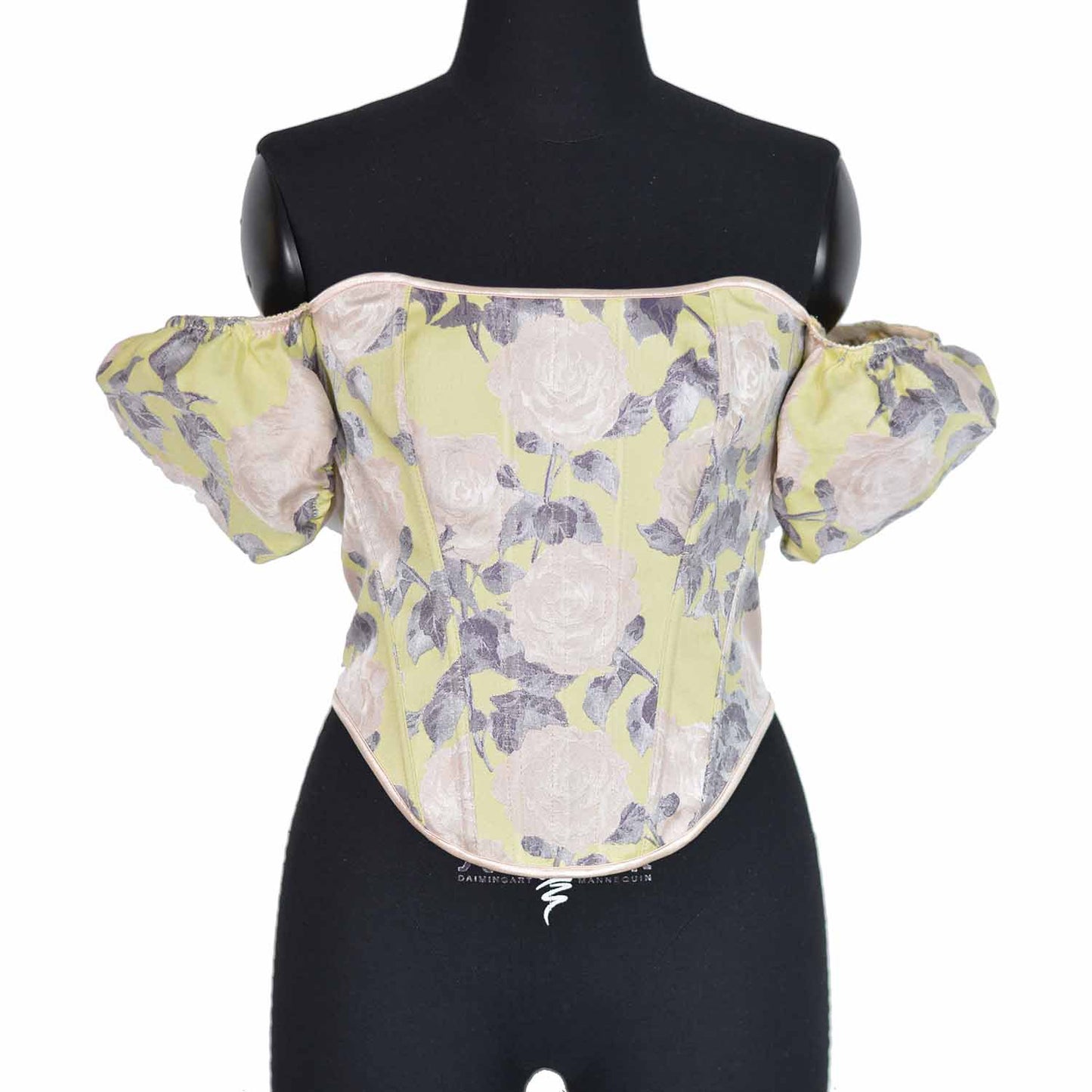 An off-the-shoulder corset top with a pale yellow-green base and a floral print of large light pink roses with muted purple-gray leaves, displayed on a black dress form mannequin against a white background.