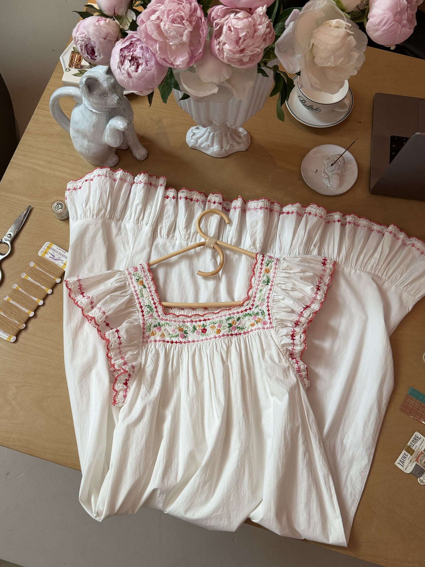 A white off-the-shoulder blouse with pink and green embroidered details, displayed on a wooden hanger on a wooden table surrounded by decorative items including pink peonies in a vase, a small gray ceramic cat figurine, and sewing notions.