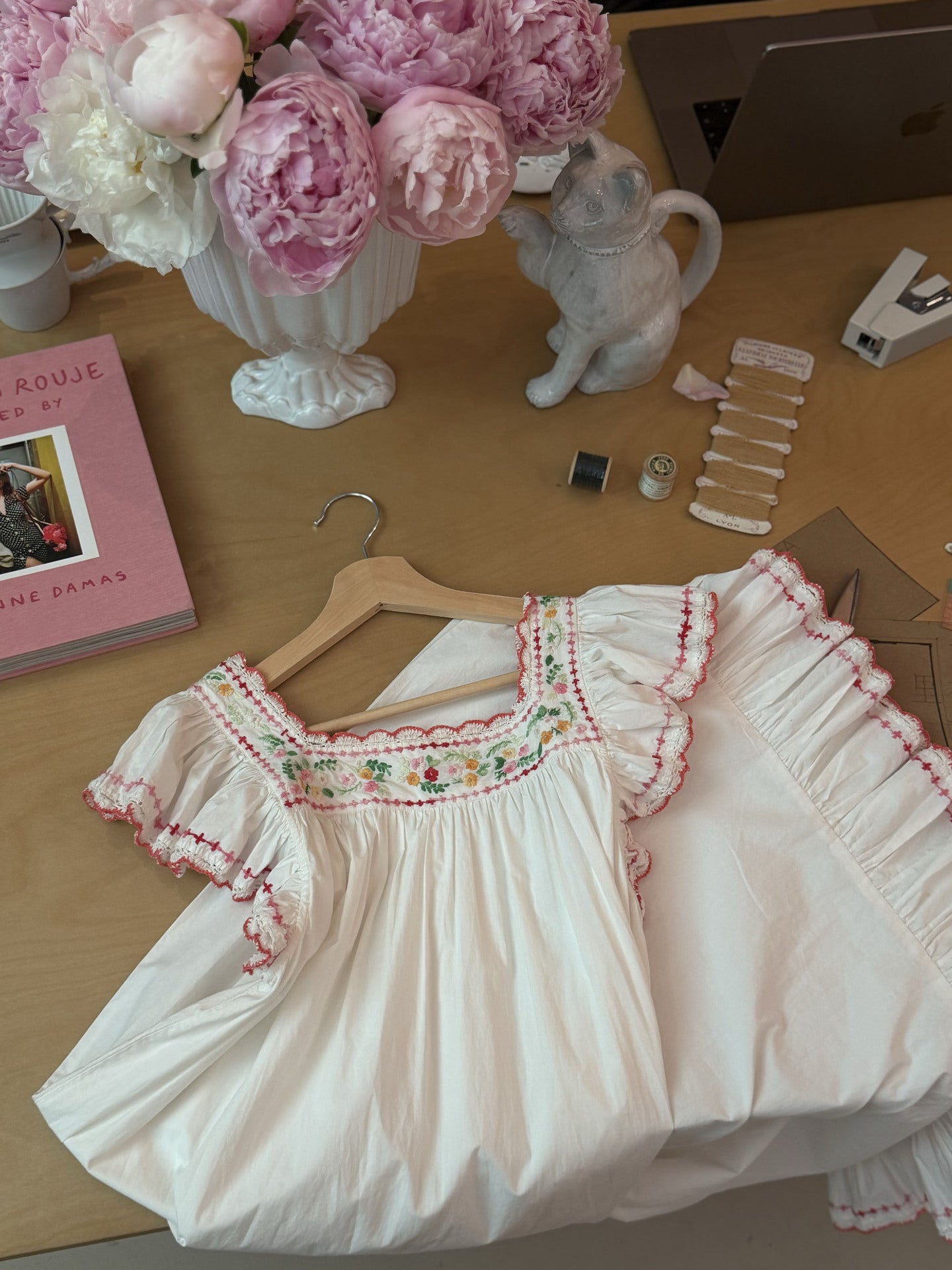 A white short-sleeve dress with pink ruffle trim and a floral embroidered yoke, displayed on a wooden hanger on a desk with flowers, a book, and small accessories around it.