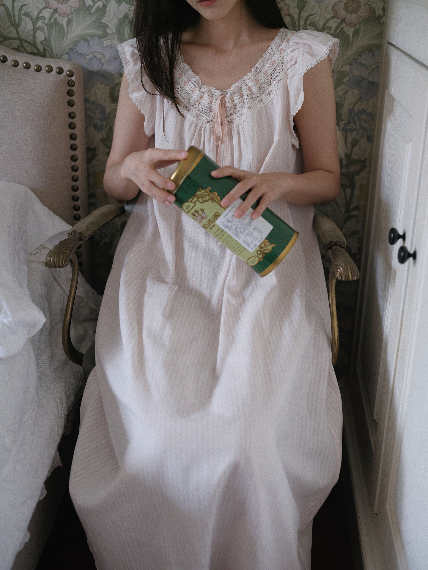 A woman wearing a white sleeveless nightgown with lace trim sits in a vintage upholstered armchair, holding a green and gold tin.