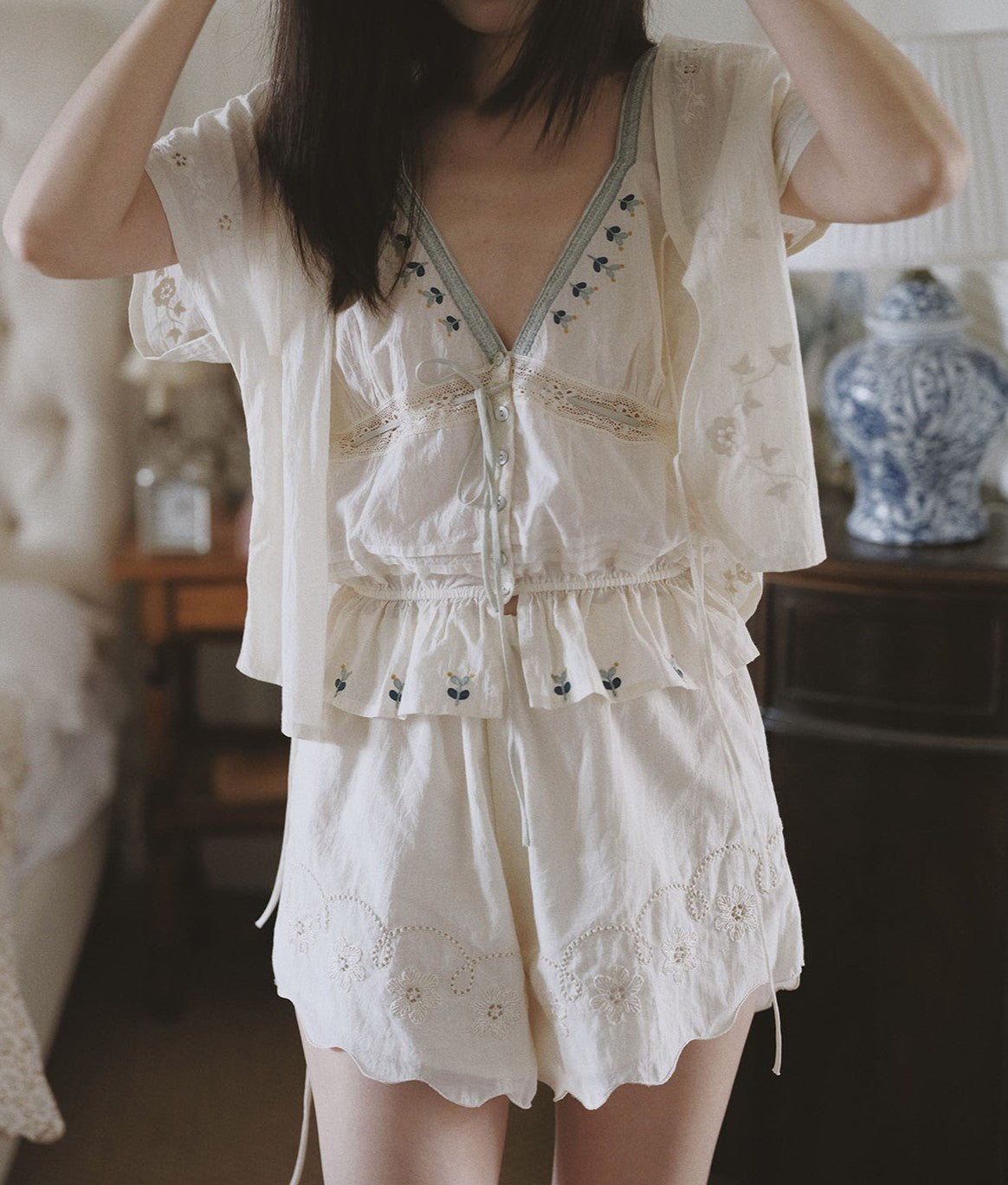 A woman wearing a cream-colored two-piece pajama set with embroidered details, featuring a short-sleeve top and matching shorts, in a softly lit bedroom setting.