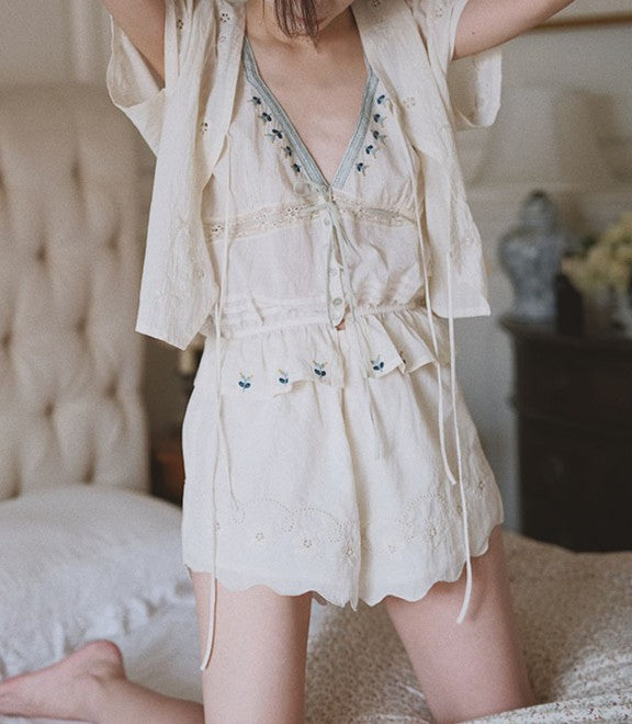 A woman wearing a cream-colored embroidered short romper with a matching tie-on vest, posed on a bed in a softly lit bedroom.