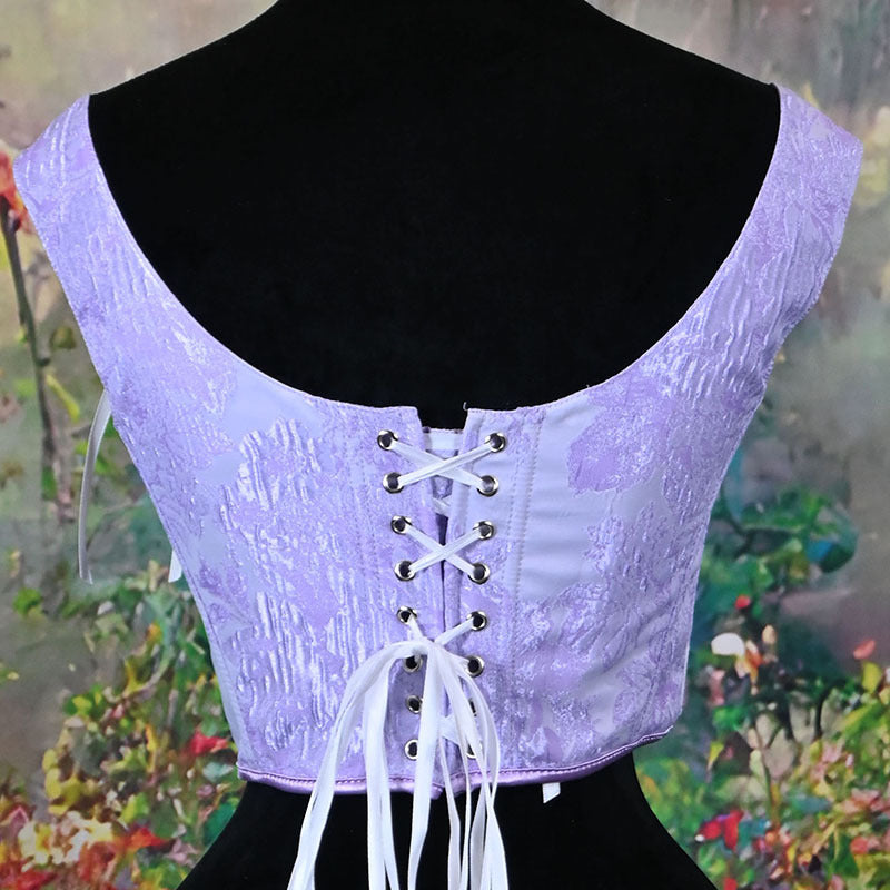 A lavender lace corset top displayed on a black mannequin torso, showing the back lace-up closure with white laces and silver eyelets against a blurred garden background.