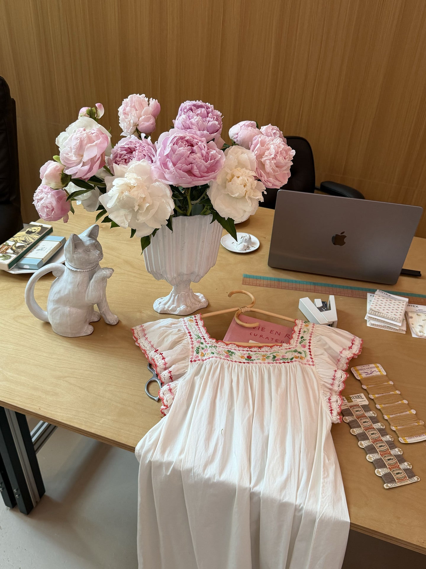 A white sleeveless dress with pink floral embroidery and ruffled trim is laid out on a wooden desk next to a vase of pink and white peonies, a cat figurine, and a laptop.