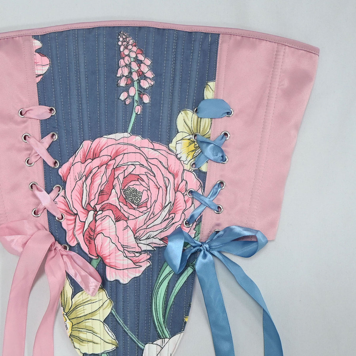 A close-up of a floral corset panel with a blue-gray striped center, pink satin side panels, pink lace-up ties, and blue satin ribbon ties, decorated with large pink flowers and yellow daisies.