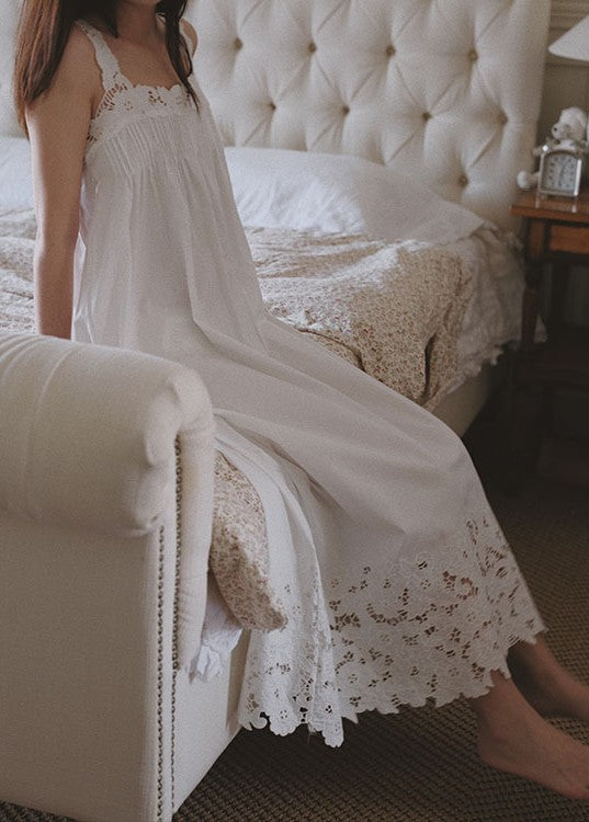 A woman wearing a white sleeveless lace nightgown sits on a cream tufted bed in a softly lit bedroom.