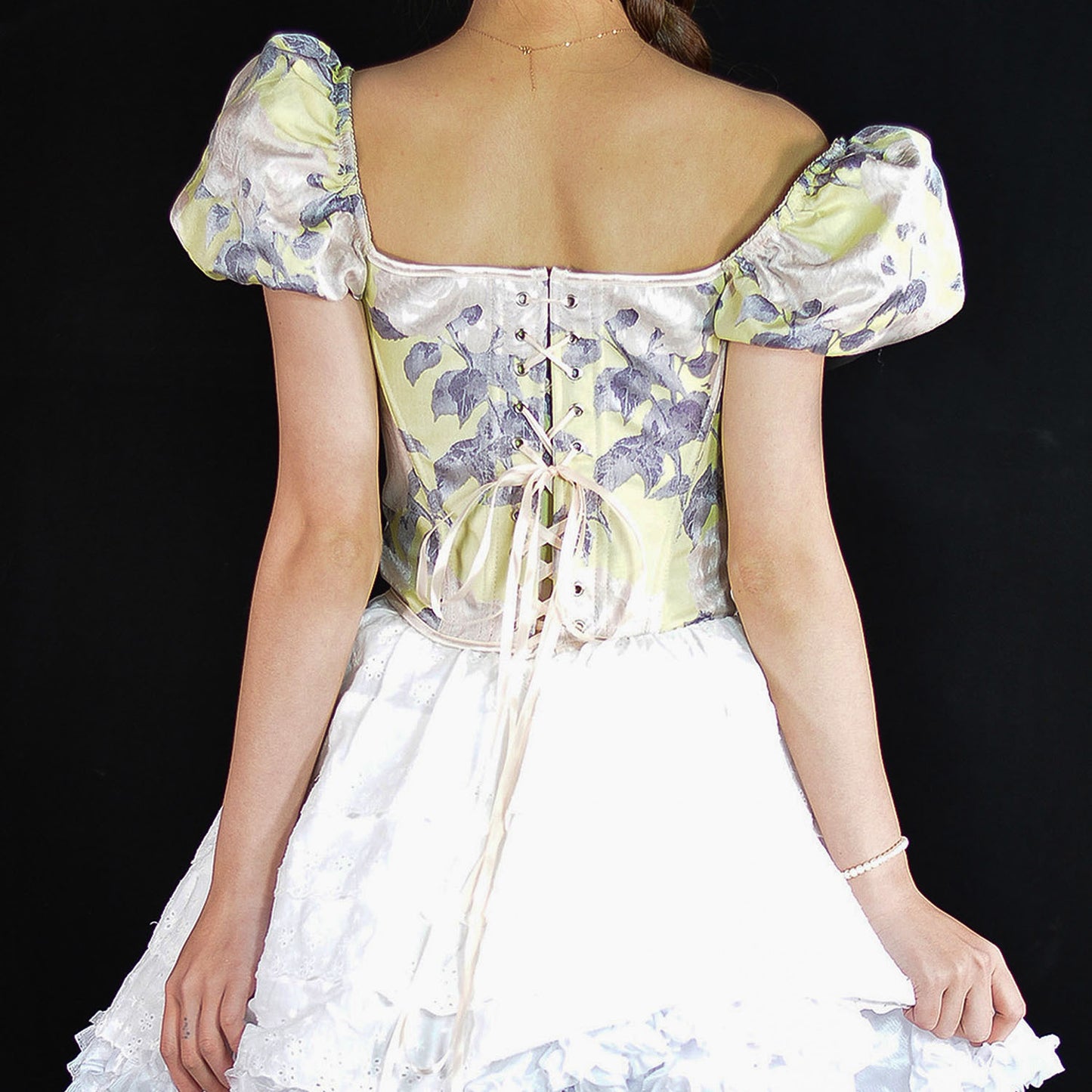 A woman shown from the back wearing a vintage-style Lolita dress with a pale yellow and lavender floral corset bodice, puffed sleeves, and a white ruffled skirt against a black background.