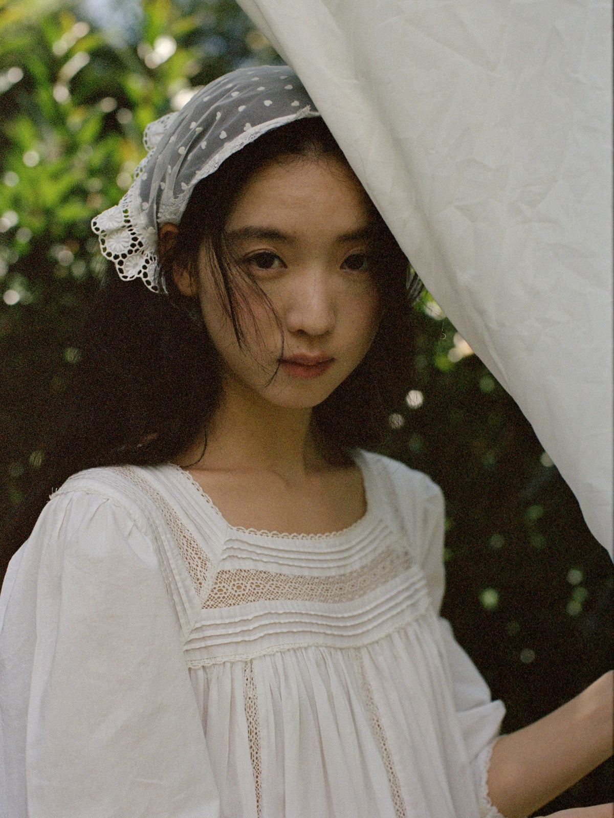 A young woman wearing a white embroidered blouse and a gray polka-dot headscarf, standing outdoors under a white fabric canopy with green foliage in the background.