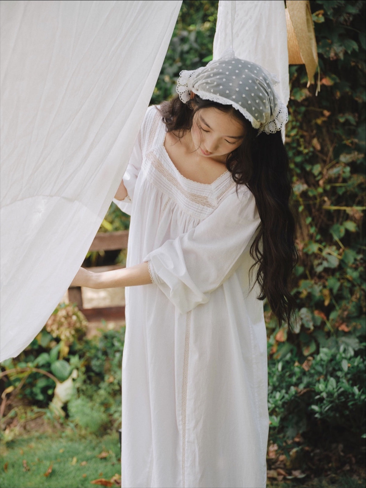 A woman in a white long-sleeve nightgown and a polka-dot bonnet stands outdoors next to a white hanging sheet, with green foliage in the background.