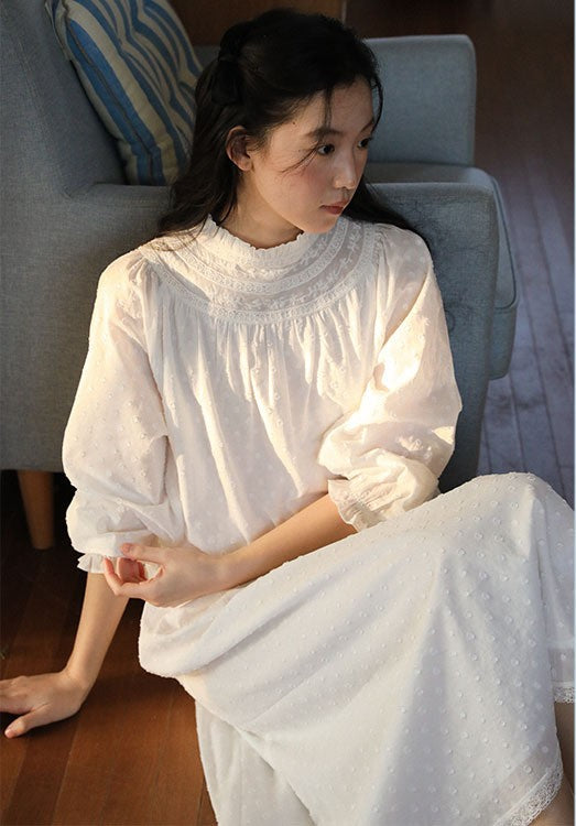 A woman sitting on a gray armchair in a cozy indoor setting, wearing a white long-sleeve dress with a ruffled high neck and eyelet fabric details.