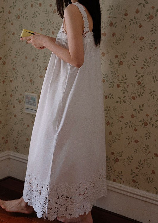 A woman wearing a long white nightgown with lace trim, standing in a room with floral wallpaper and bare feet on a wooden floor.