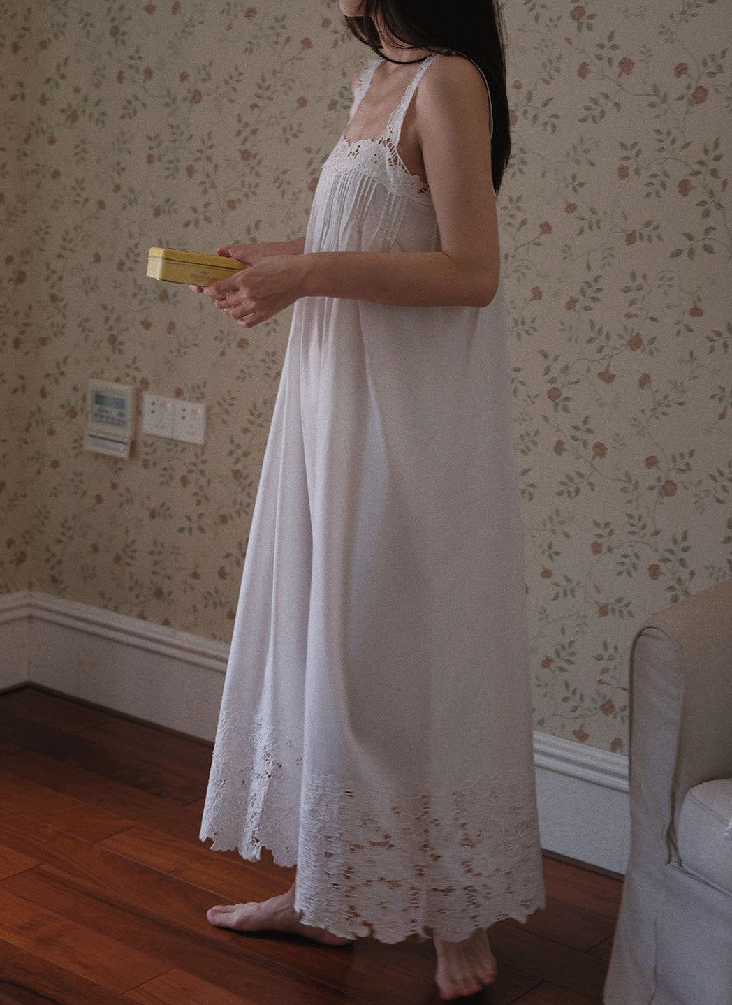 A woman wearing a long white nightgown with lace trim, standing barefoot in a room with floral wallpaper and a wooden floor.