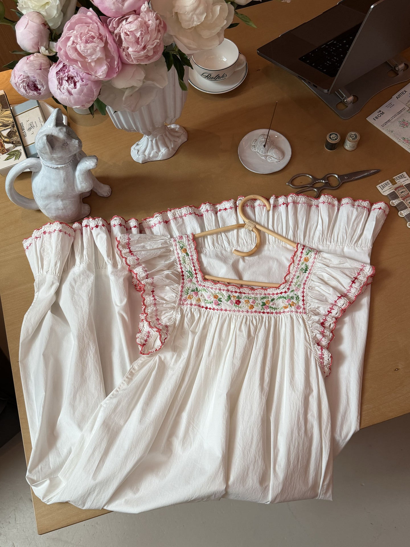 An off-white cotton smocked blouse with pink ruffle trim and colorful floral embroidery on the chest, laid flat on a wooden desk surrounded by sewing and floral props.