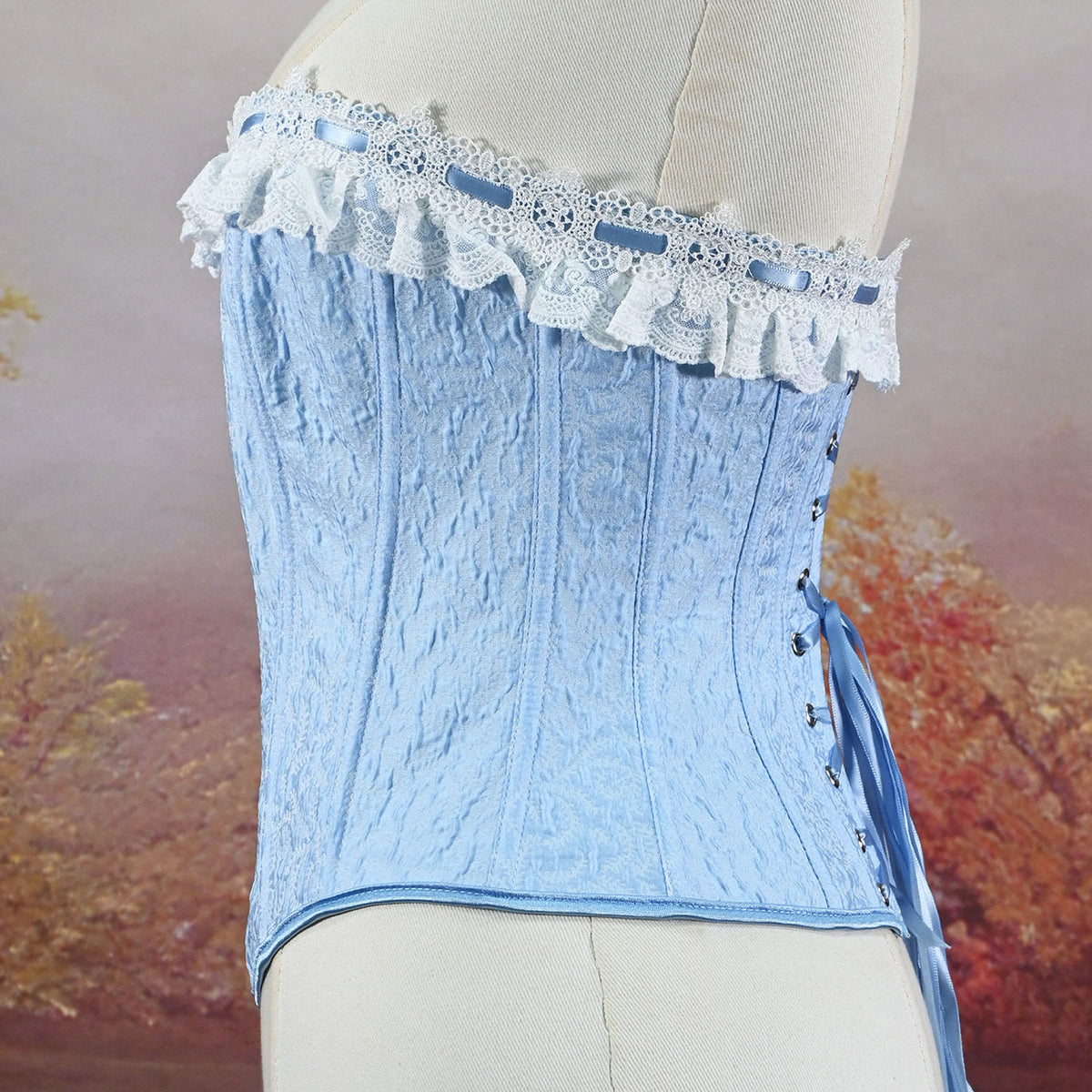 A light blue lace and textured fabric corset top displayed on a cream dress form, featuring a ruffled lace band with blue ribbons and side lacing with a blue tie.