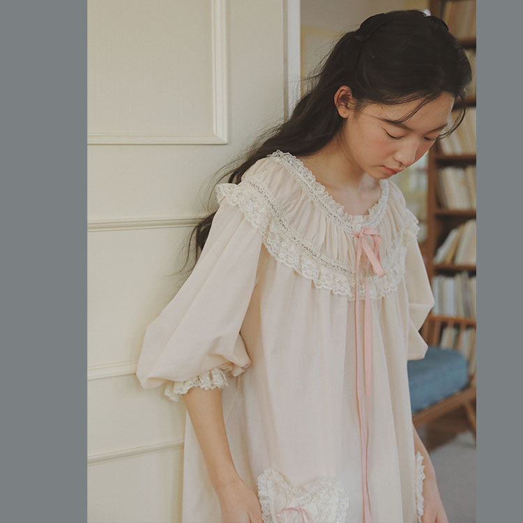 A young woman wearing a light cream-colored nightgown with white lace trim and a pink ribbon tie, standing in a softly lit indoor setting.