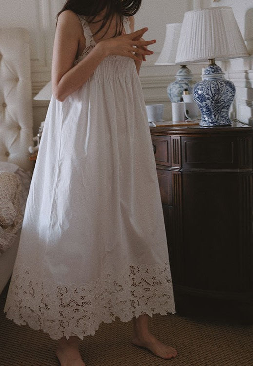 A woman wearing a long white cotton nightgown with lace trim at the hem, standing barefoot in a bedroom next to a dark wooden nightstand with lamps.