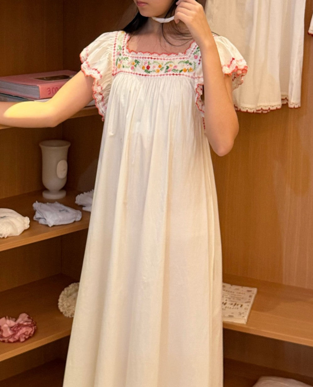A woman wearing a long white nightgown with colorful floral embroidery around the neckline and ruffled sleeves, standing in front of a wooden shelf display.