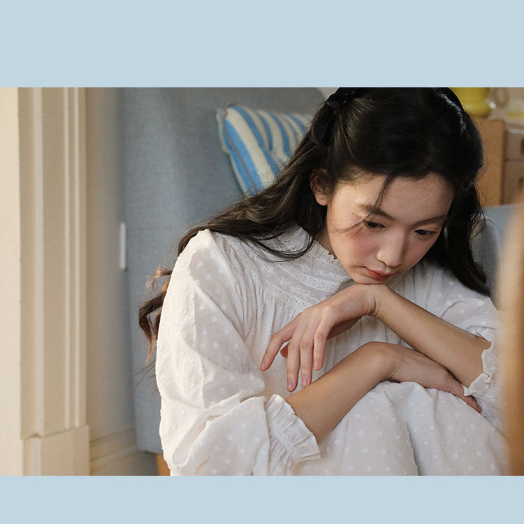 A young girl with long dark hair sits curled up indoors, wearing a white long-sleeve top with small polka dots. The photo is taken in a softly lit room with a striped cushion and light-colored furniture in the background.
