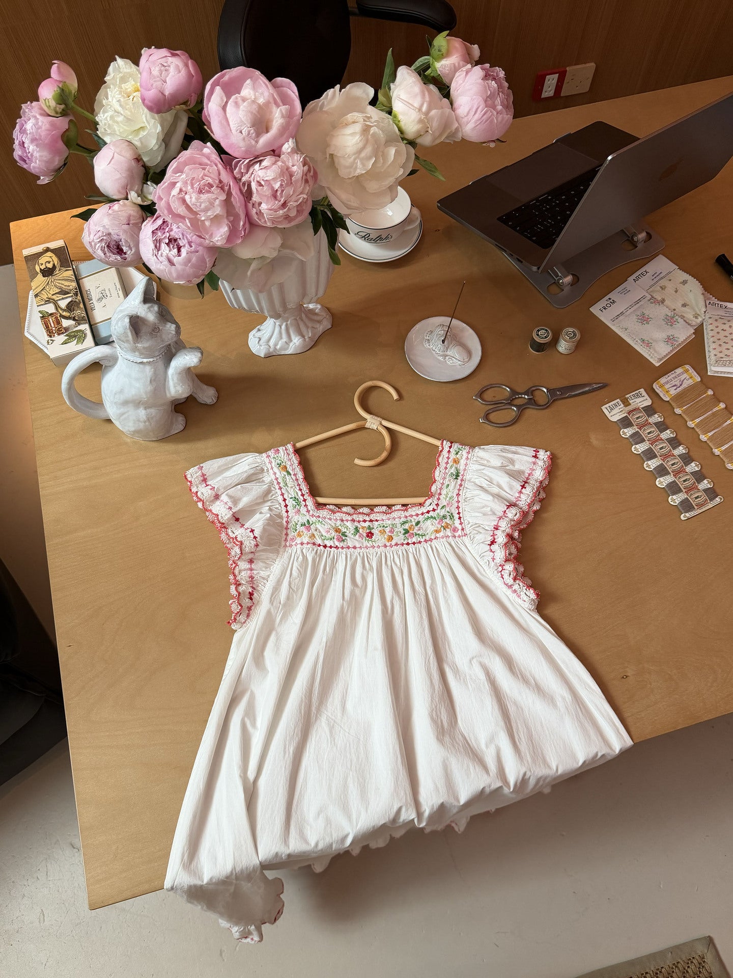 A white baby dress with pink ruffled trim and colorful embroidered details on the chest, displayed on a wooden table with a wooden hanger. The background features a bouquet of pink and white peonies, a white ceramic cat figurine, scissors, and other craft or stationery items.