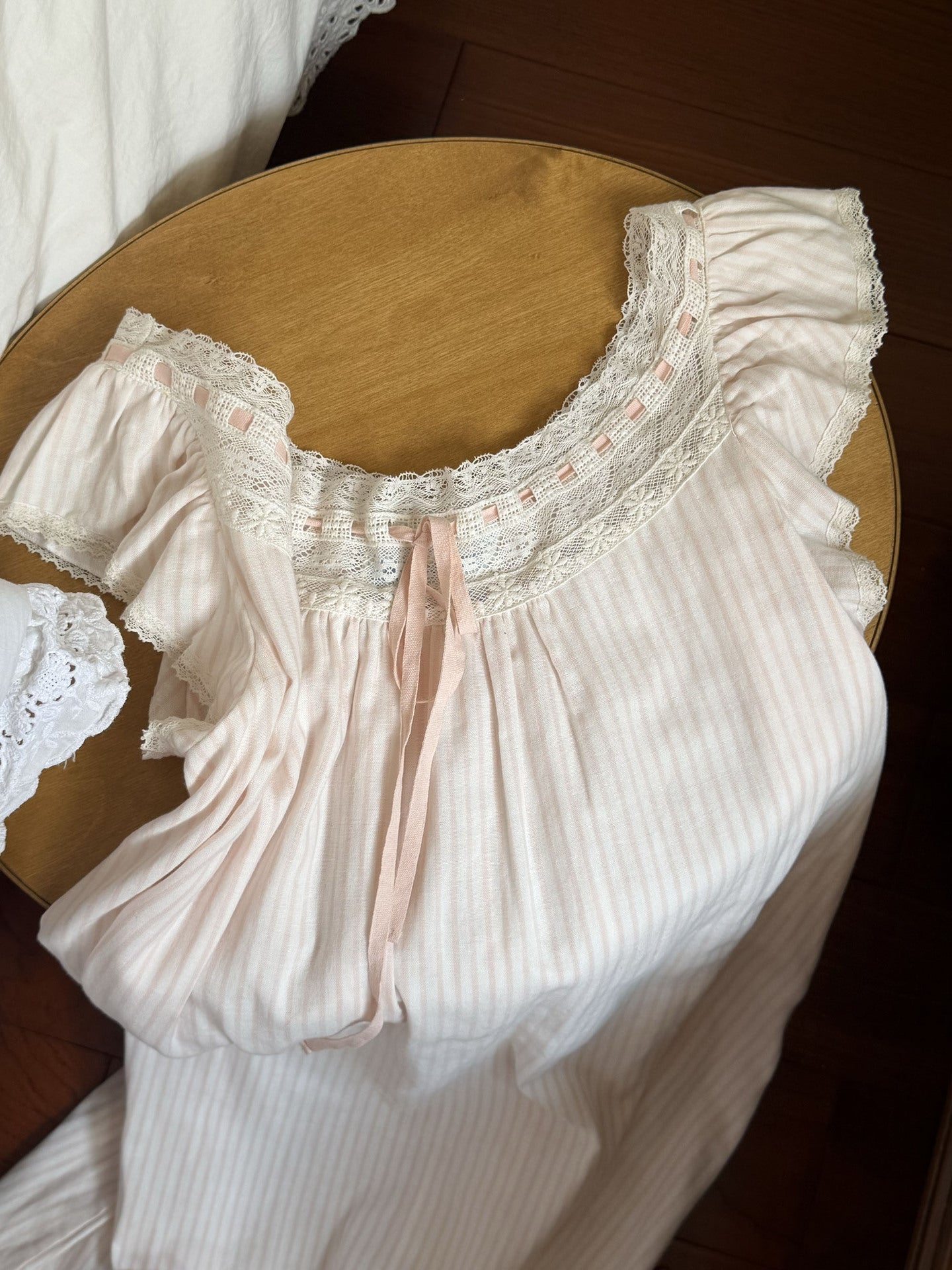 A pale pink and white vertically striped nightgown with ruffled sleeves and a lace neckline, laid flat on a round wooden surface.
