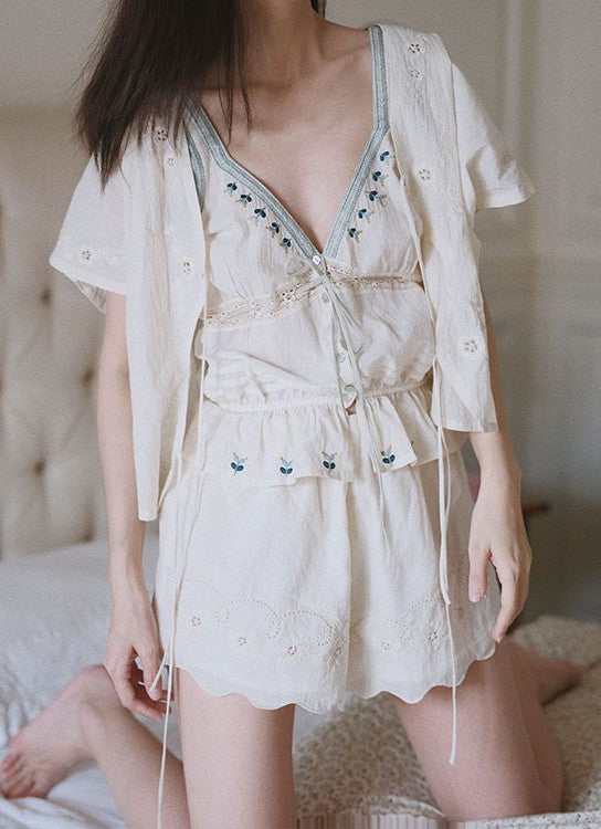 A woman wearing a light cream-colored two-piece loungewear set consisting of a short-sleeve button-up shirt and matching ruffled shorts with delicate floral embroidery, posed in a softly lit bedroom setting.