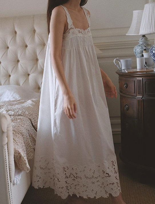 A woman wearing a long white nightgown with lace trim, standing in a bedroom next to a tufted bed and a wooden nightstand with a lamp.
