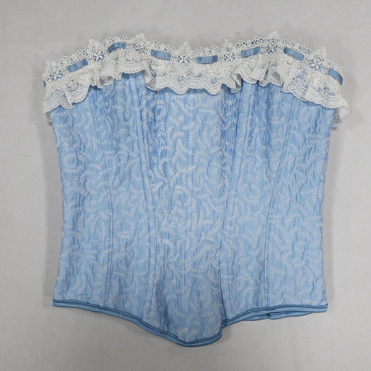 A light blue lace corset with white floral lace trim and ruffles along the waistband, laid flat on a neutral gray background.