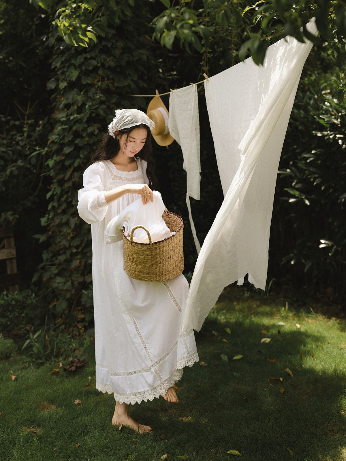 A woman in a long white dress stands barefoot on a grassy lawn, holding a woven basket with folded white fabric. A string line with more white garments hangs in the background among leafy green trees.