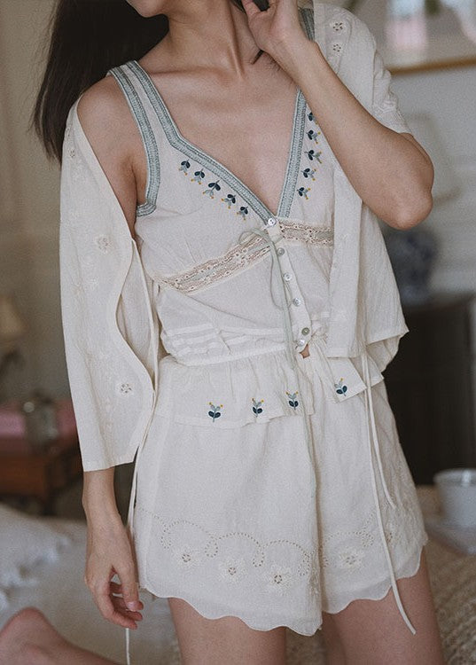 A woman wearing a cream-colored embroidered romper with a matching long-sleeve cardigan. The outfit features delicate floral embroidery, a V-neckline, and a button-up front, shown in a softly lit indoor setting.