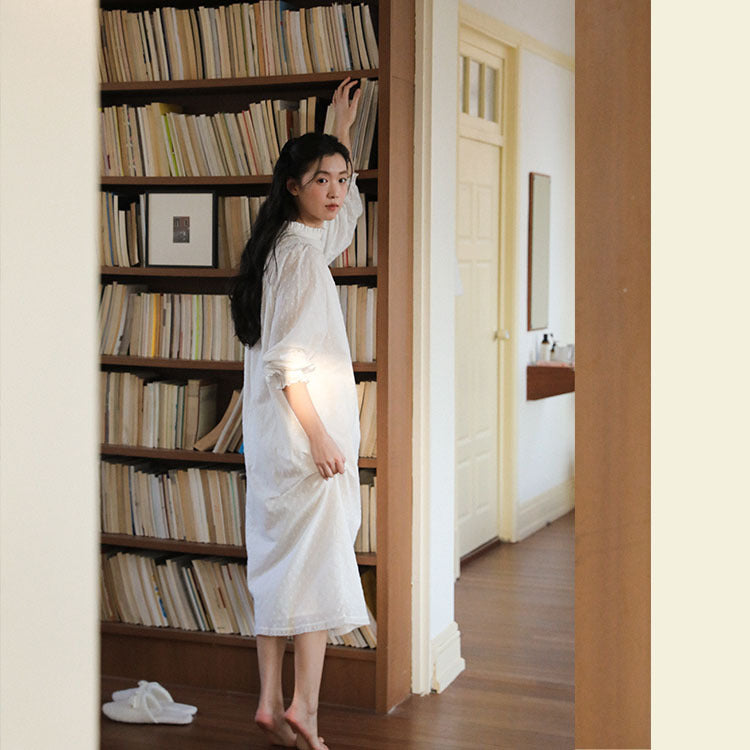 A woman stands barefoot in a doorway next to a tall wooden bookshelf filled with books, wearing a loose white long-sleeve dress. A white slipper is visible on the floor in the lower left.