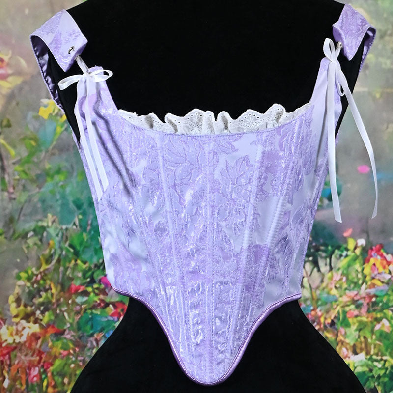 A lavender lace corset top displayed on a black dress form, featuring floral lace panels, white ruffled trim, and white tie straps, with a blurred garden background.