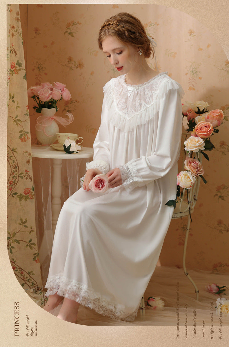 A woman wearing a long white satin nightgown with lace trim sits in a vintage-style room with floral wallpaper and pink roses.