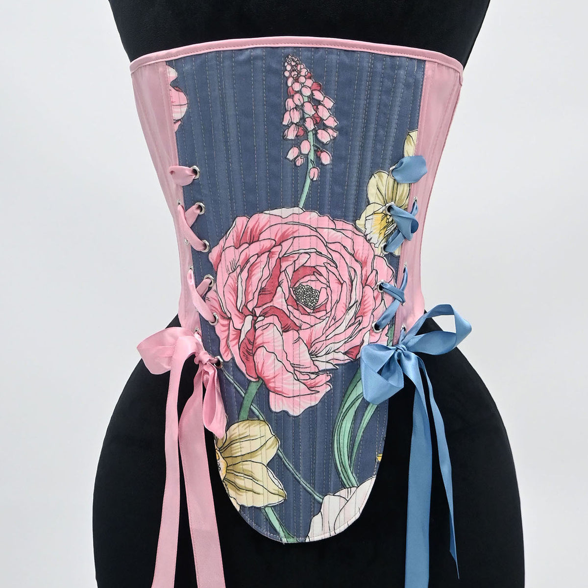 A corset-style waist trainer displayed on a black mannequin torso against a light gray studio background. The garment has a dark blue-gray striped base with a large pink floral print, pink trim, lace-up side details, and pink and blue ribbon ties.
