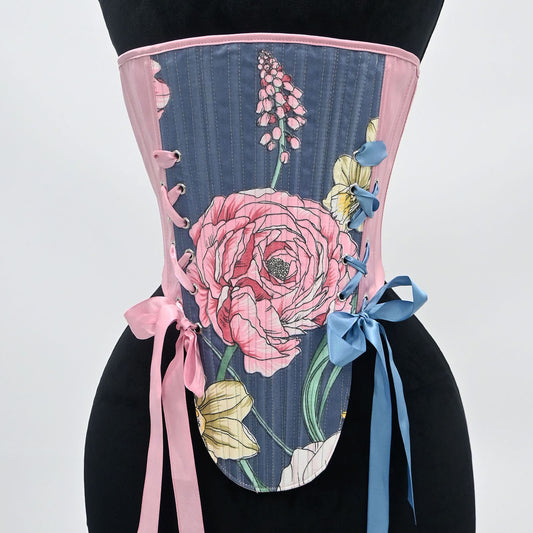 A corset-style waist trainer displayed on a black mannequin torso against a light gray studio background. The garment has a dark blue-gray striped base with a large pink floral print, pink trim, lace-up side details, and pink and blue ribbon ties.