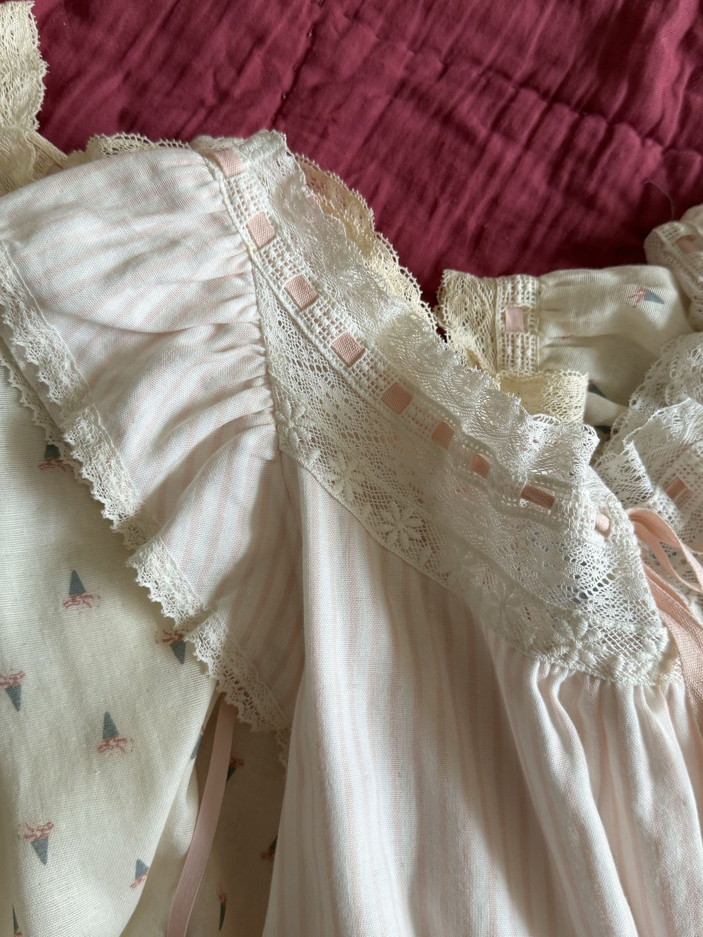 A close-up of a cream-colored ruffled blouse with delicate lace trim and small pink ribbon accents, laid flat on a dark pink textured fabric background.