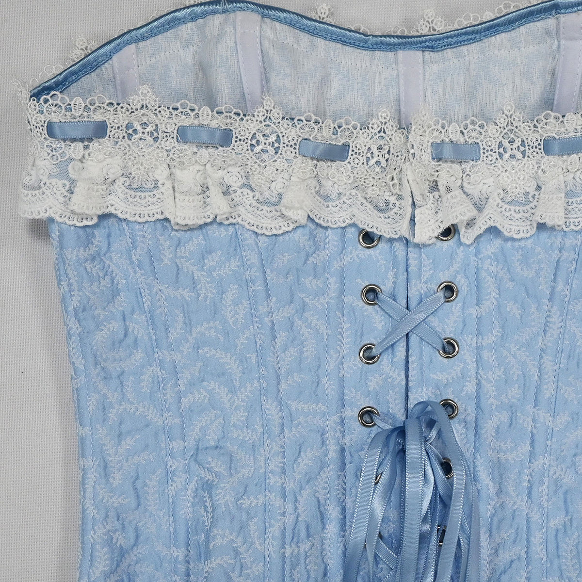 A close-up of a light blue lace-up corset with a textured floral pattern, featuring white lace trim with blue ribbon accents at the bust.