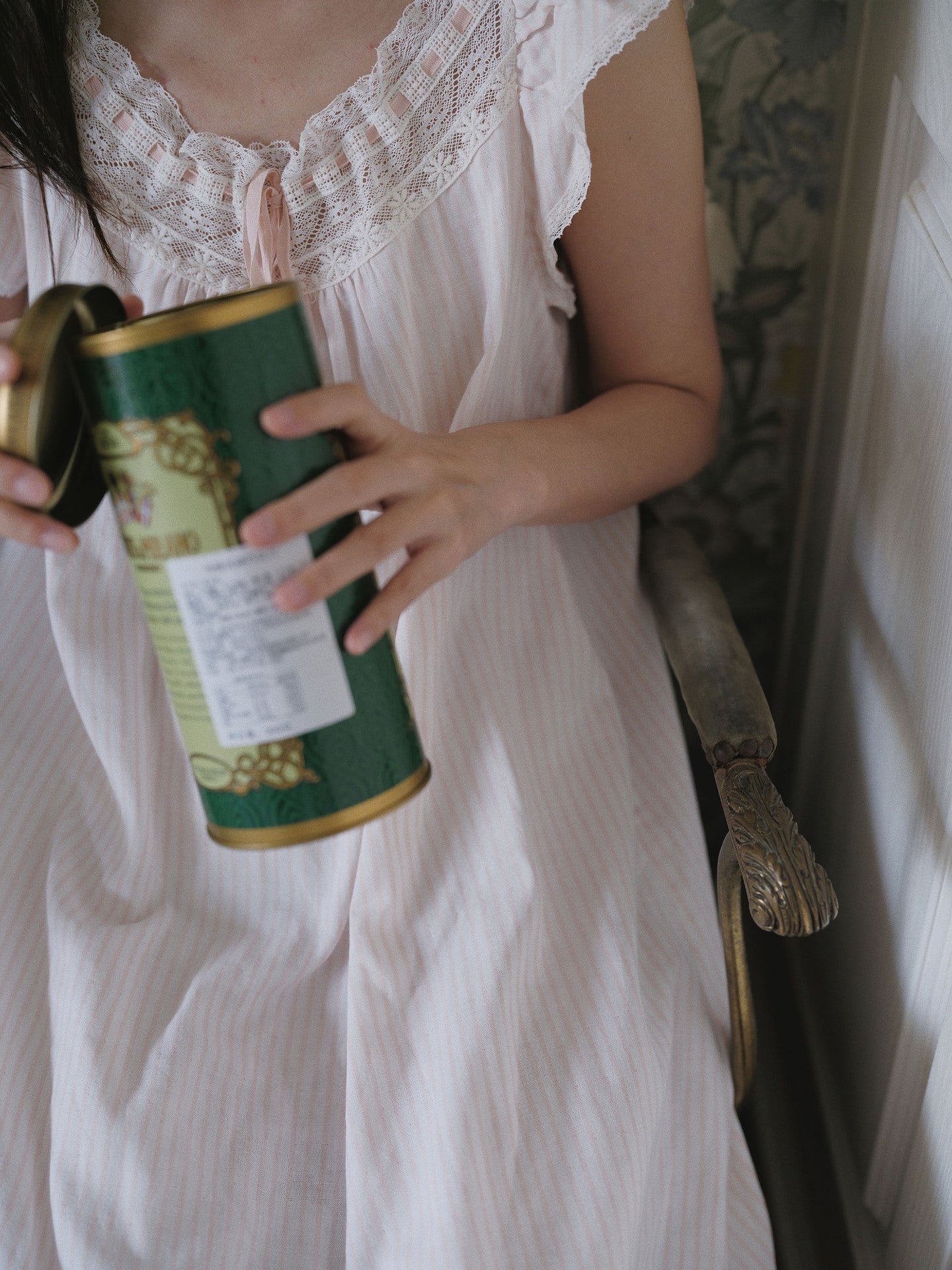 A person in a white lace-trimmed nightgown holds a green cylindrical tin with gold trim and a white label in a softly lit indoor setting.