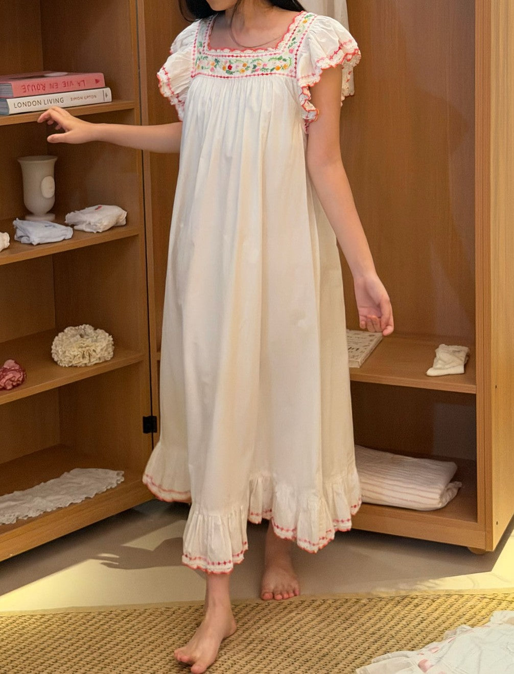 A woman wearing a long white nightgown with colorful floral embroidery on the chest and pink ruffled trim, standing barefoot in a bedroom next to a wooden shelf.