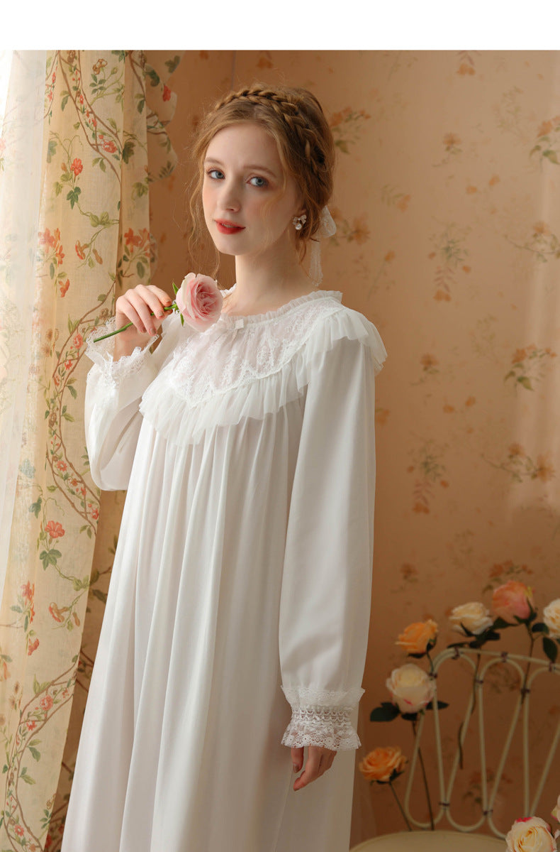 A woman wearing a long white nightgown with lace trim and ruffled details, standing in a vintage-style room with floral wallpaper and curtains.