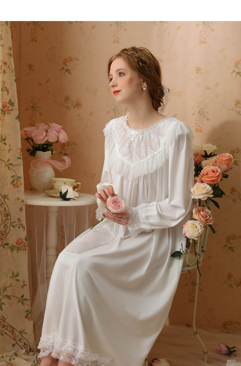 A woman wearing a long-sleeved white satin nightgown with lace trim, sitting in a vintage-style room with floral wallpaper and pink roses.