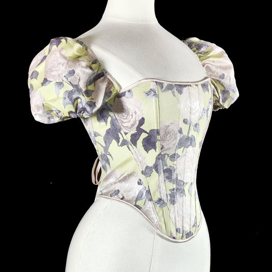 A vintage-style corset top with puff sleeves, displayed on a white mannequin torso against a solid black background. The fabric features a floral pattern in pale sage green, lavender, and white.
