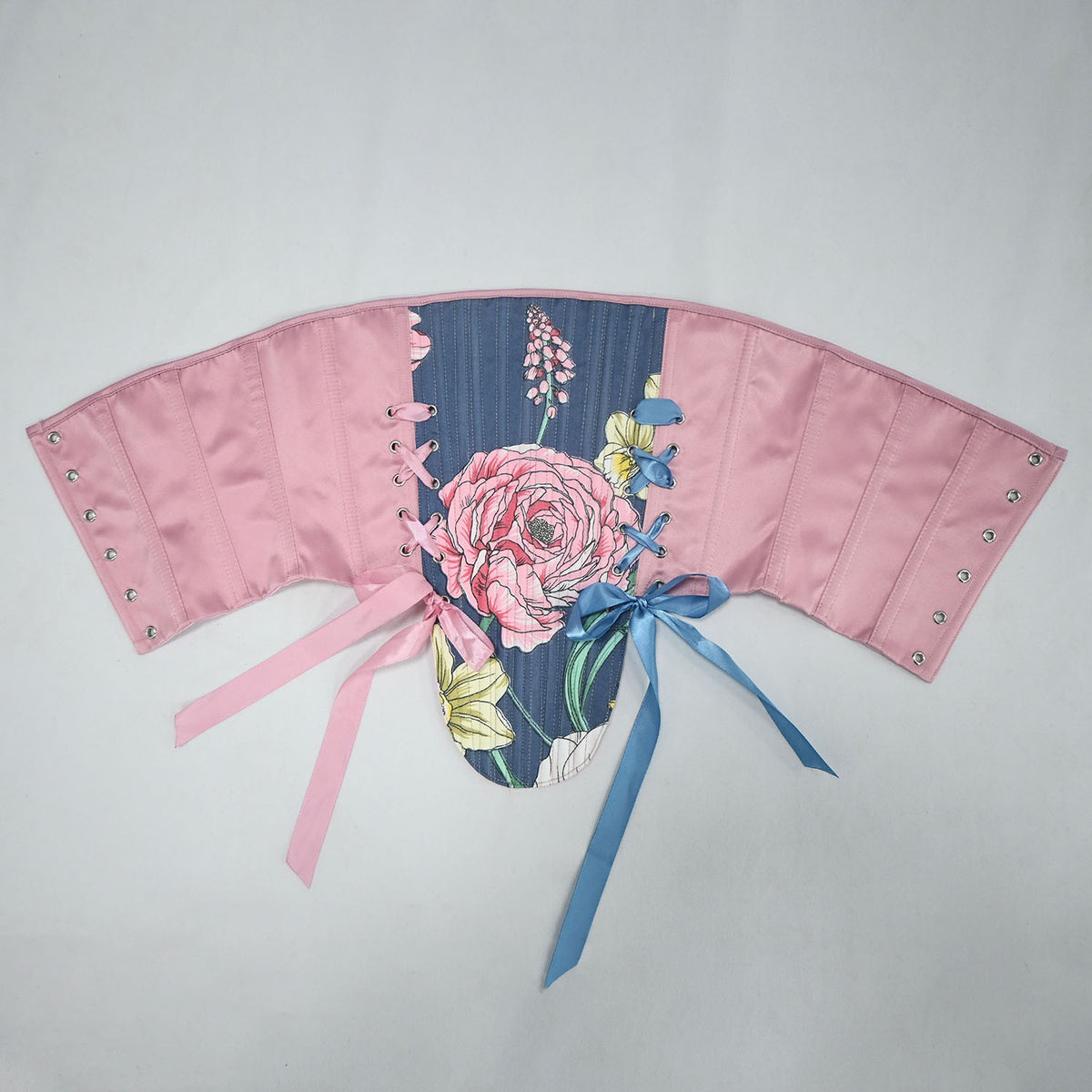 A pink fabric corset-style accessory with floral printed panels, side lacing, metal eyelets, and two ribbon ties in pink and blue, laid flat on a light gray background.
