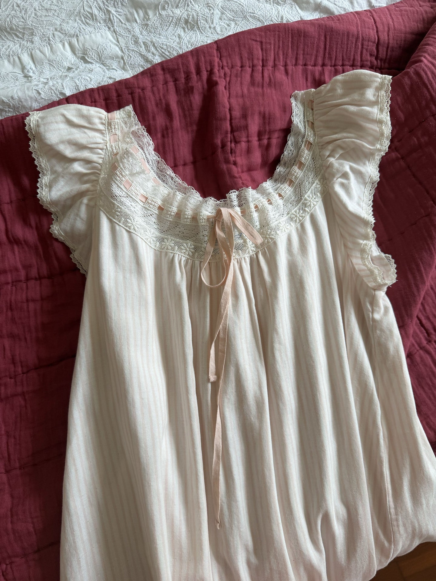 An off-white cotton nightgown with ruffled sleeves, a lace-trimmed scoop neckline, and a center tie, laid flat on a dark red quilted blanket.