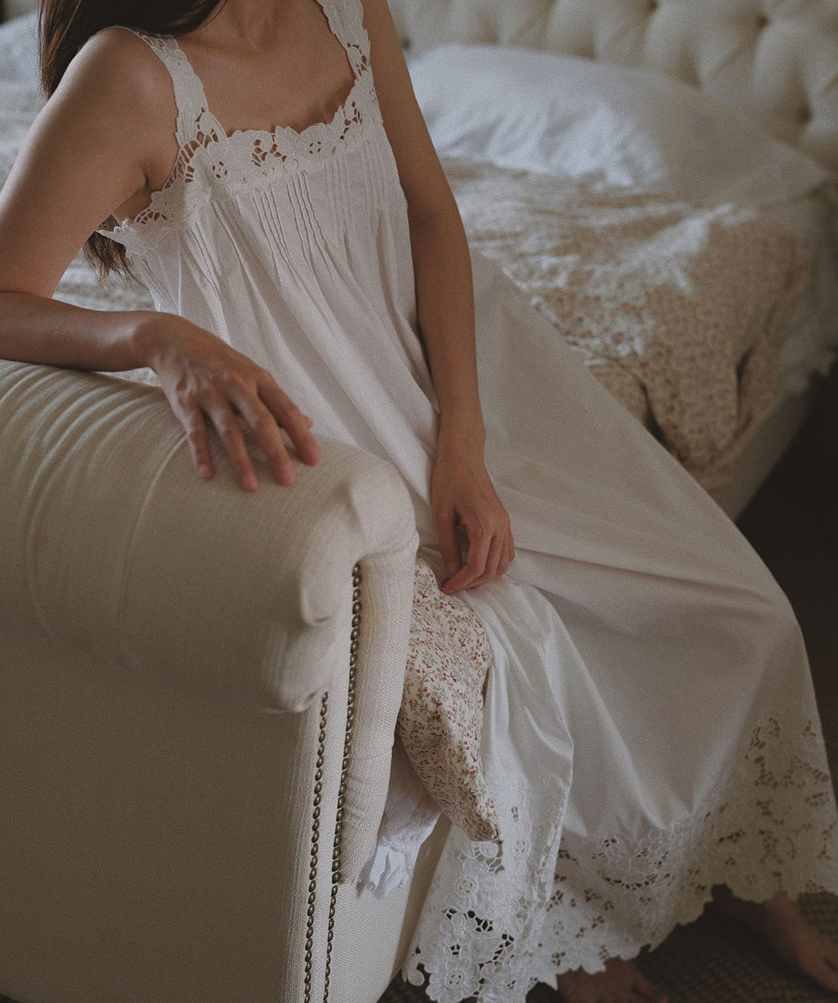 A woman wearing a white nightgown with delicate lace trim, sitting on a cream-colored upholstered armchair in a softly lit bedroom.