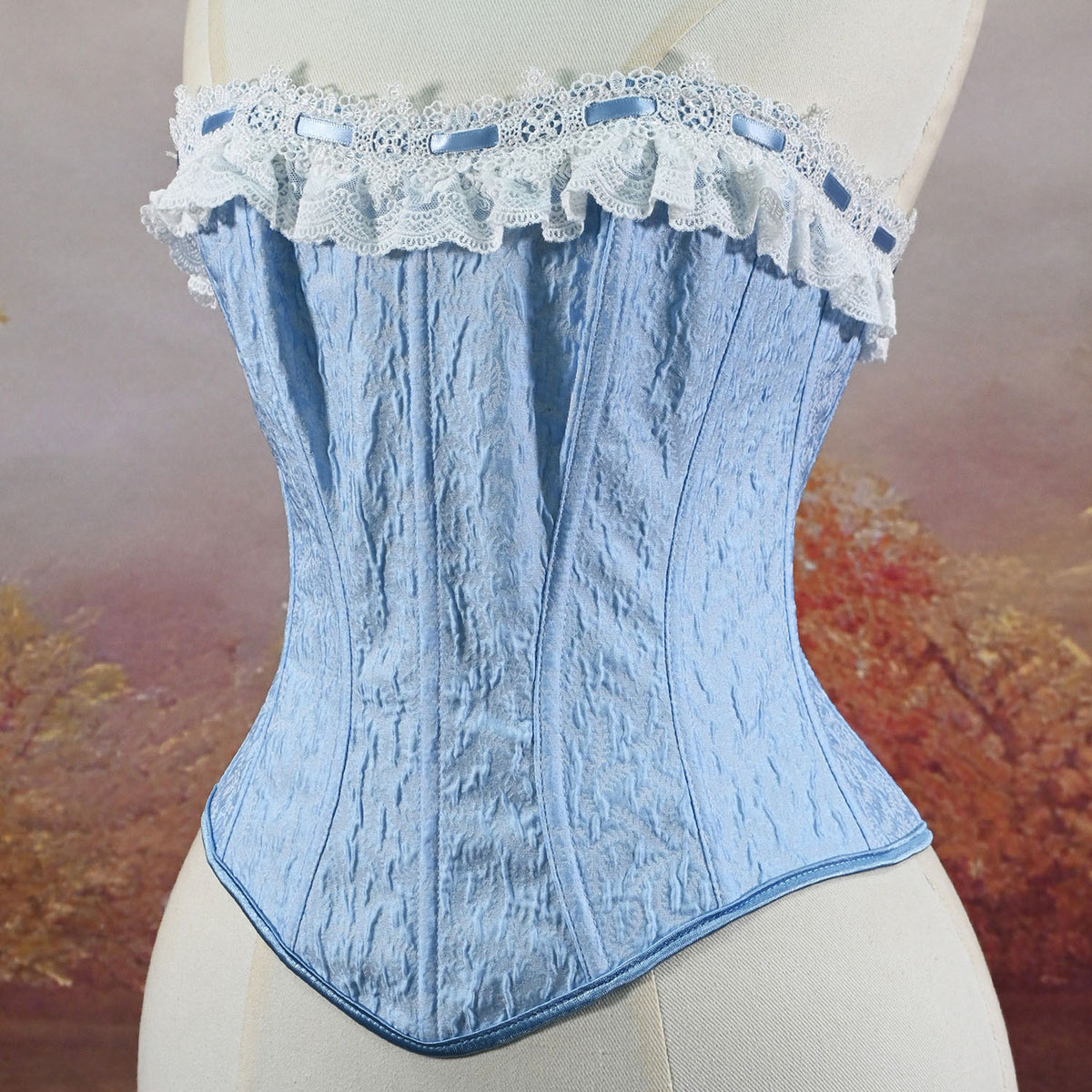 A light blue lace and fabric corset bustier displayed on a cream-colored dress form mannequin, featuring a ruffled lace top band with blue ribbon accents and a textured corset body.